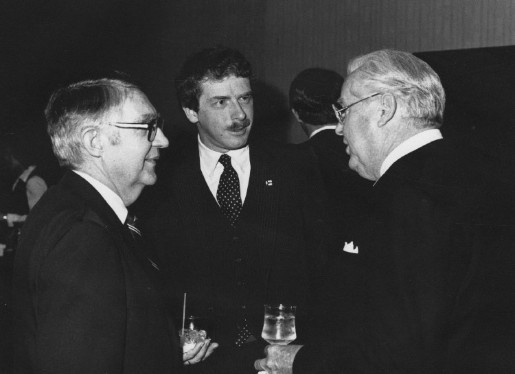 White man with mustache in suit and tie standing in between two older white men in suits with glasses talking