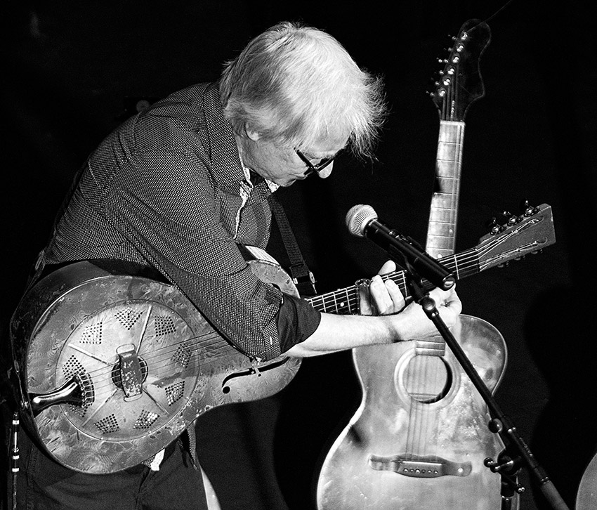 Older white man on stage in front of microphone with two guitar-shaped instruments