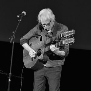 Older white man standing on stage at microphone with a two-necked guitar