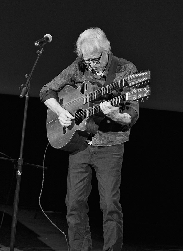 Older white man standing on stage at microphone with a two-necked guitar