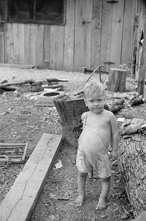 White child standing with logs and house behind him
