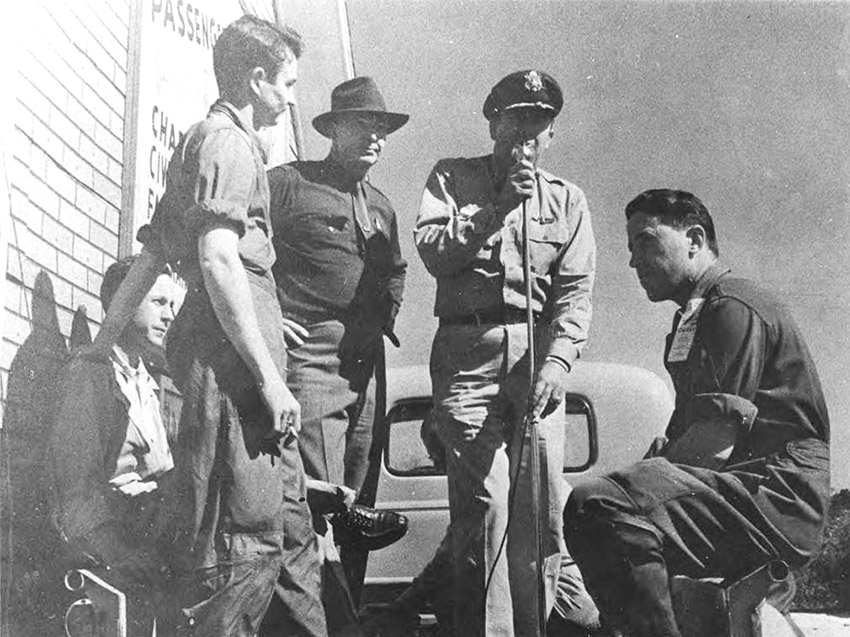 Group of white men, one in military uniform speaking into microphone
