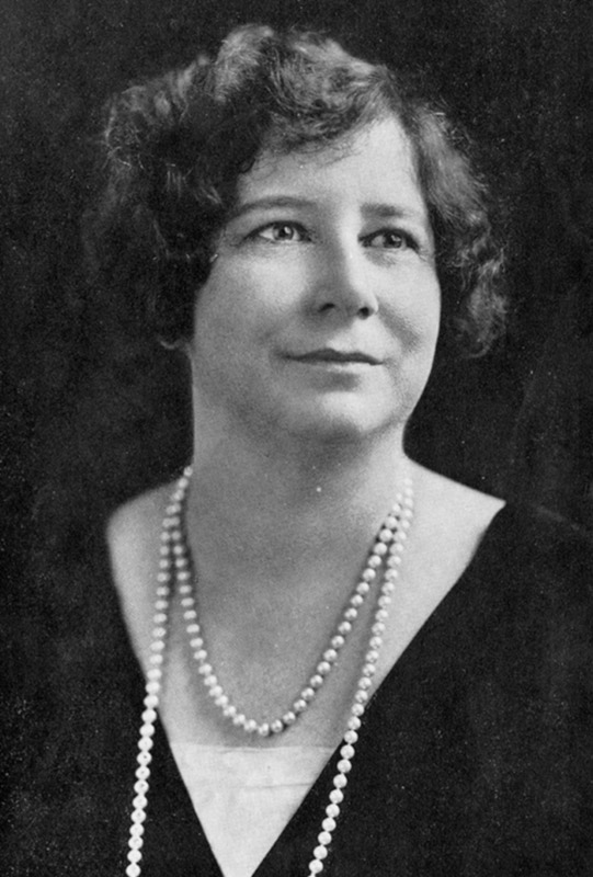 White woman with curly hair smiling in dress and two strings of pearls