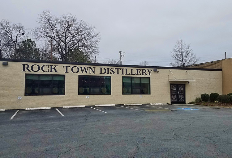 Single-story brick storefront with "Rock Town Distillery" painted above three large windows and parking lot