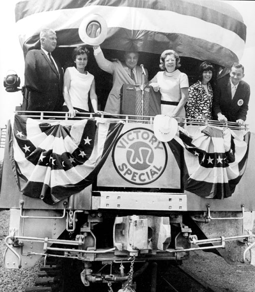 White men and women on the back of a train car labeled "W R Victory Special" with man in center standing in front of  a microphone and raising a cowboy hat in his right hand.