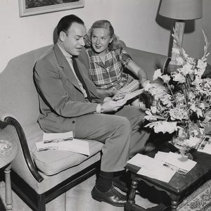 White man in suit and white woman in plaid shirt sitting together on couch reading papers