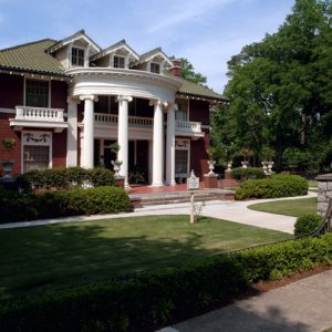 Multistory house with round covered porch supported by columns on grass with decorative bushes and sidewalk