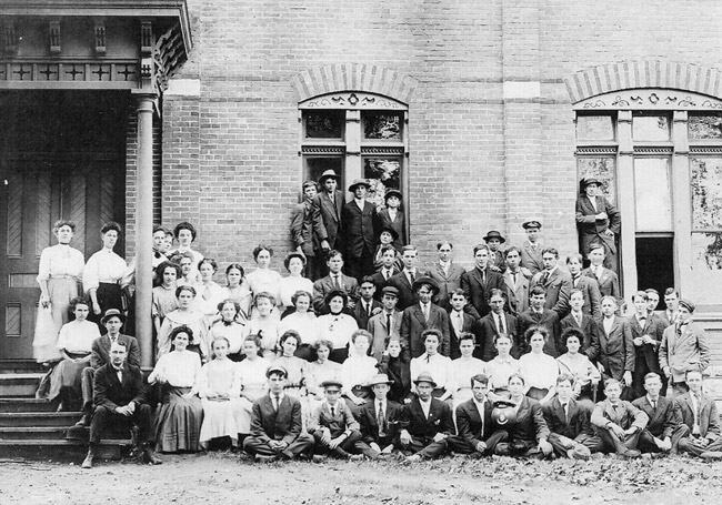 White male and female students and teachers pose for a group photo outside brick building with arched windows and covered entrance