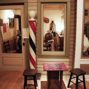 Exhibition display with two stools and checkerboard on crate with barber's pole outside barbershop display with barber's chair and pot-bellied stove