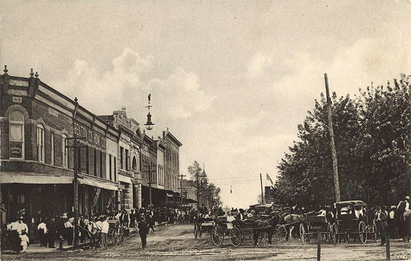 People and horse drawn carriages on crowded town street with multistory brick buildings on one side
