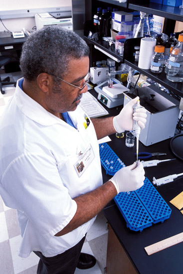 African-American man with glasses and white short-sleeved lab coat working in a chemistry lab using a pipette