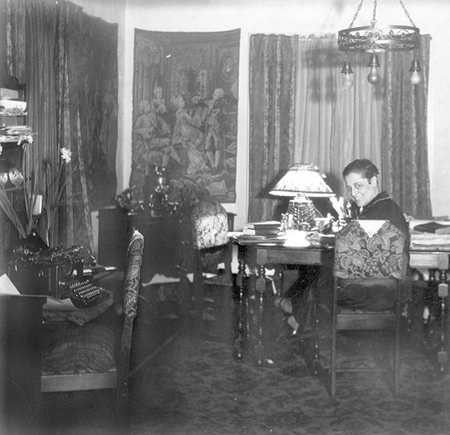 White woman sitting at table in room with hanging light fixture and desk with typewriter