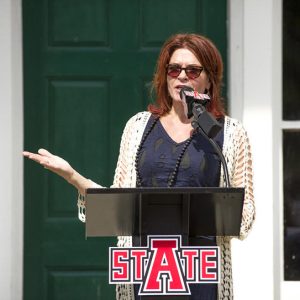 White woman with red hair speaking into a microphone at a lectern