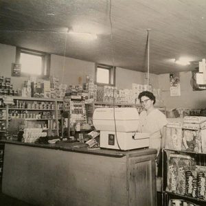 White woman with glasses sitting behind the counter in general store