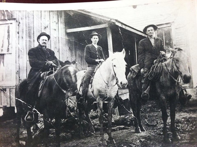 Three white men on horseback outside single-story building with covered porch