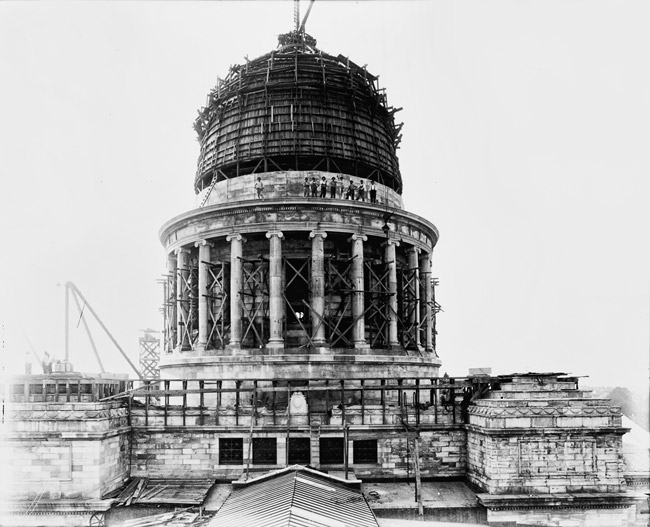 Unfinished dome with cranes and workers standing on a ledge above its columns