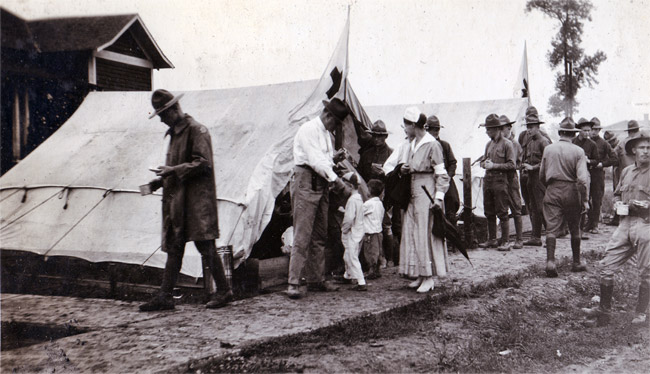 Group of white soldiers in uniform and nurses at camp with row of white tents and children