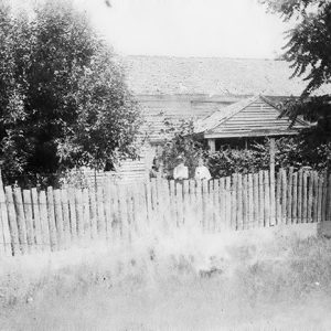 White man and woman standing outside single-story house inside wooden fence
