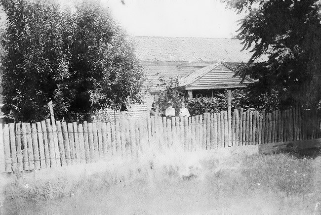 White man and woman standing outside single-story house inside wooden fence