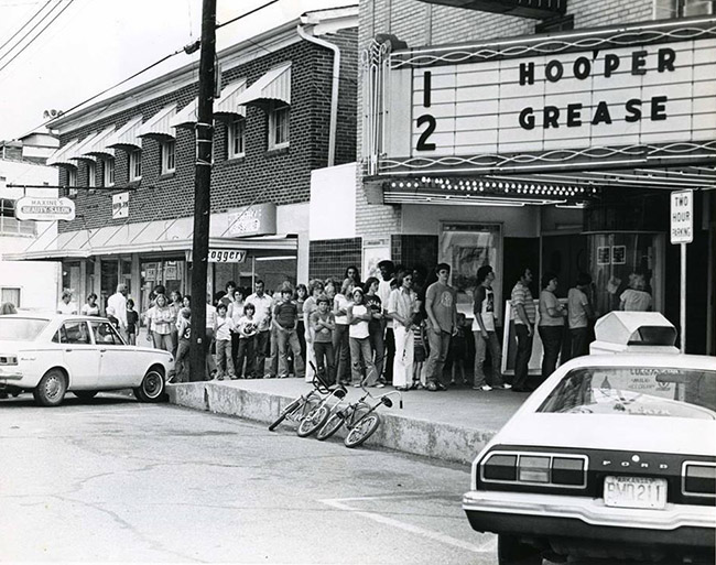 Mixed crowd of people in line outside multistory brick building and theater with "Hooper" and "Grease" on its marquee