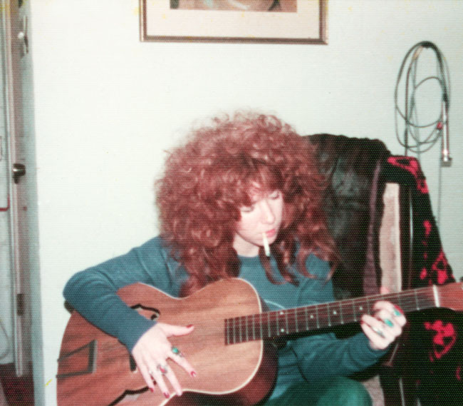 White woman with red hair playing guitar and smoking a cigarette indoors