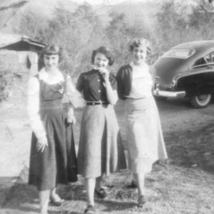 Three smiling white women in skirts standing with car behind them
