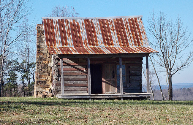Abandoned log cabin with rusted metal roof and covered porch and stone chimney