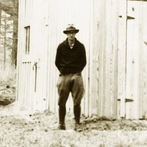 white man in hat and outdoor clothing stands beside wooden structure