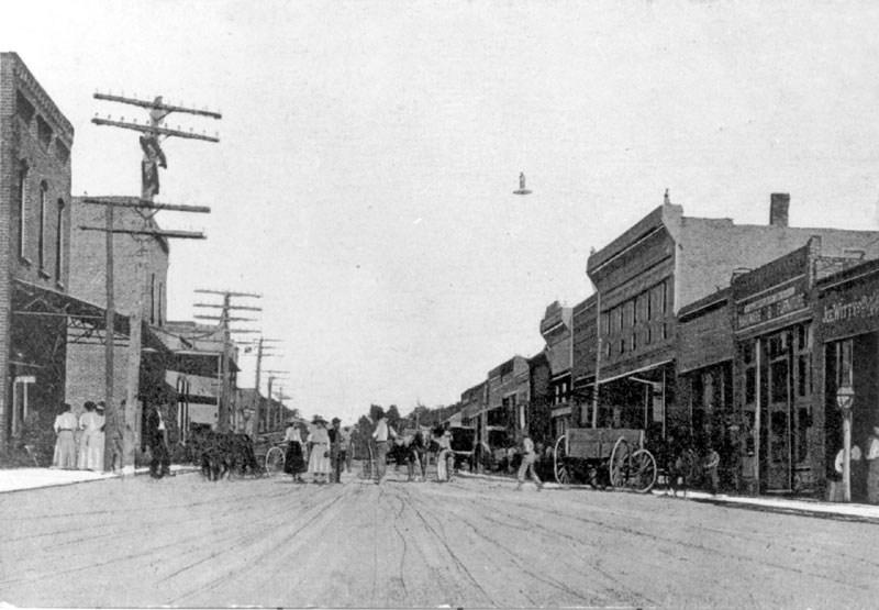 Street lined with buildings and telephone poles with crowd of people and wagons