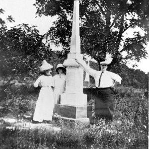 Two white women and white man posing with a tall marble monument surrounded by trees