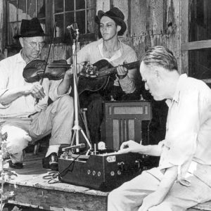 Old white man kneeling at a recording device playing a fiddle with a young white man sitting playing guitar next to him while another monitors the recording equipment