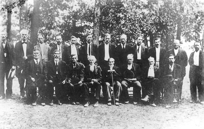 Group of old white men in suits sitting and standing with trees in the background