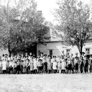 Crowd of white children outside two-story wooden school house