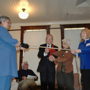 Older white man and woman cutting a ribbon being held up by white women