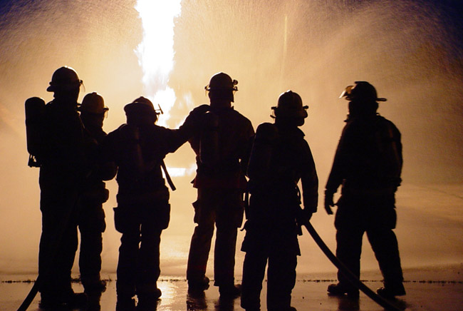 Six firefighters stand spraying hose silhouetted in front of fire at night