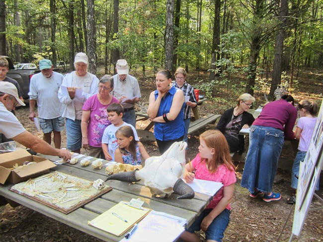 White man teaching a group of white men women and children at outdoor table with animal bones on it