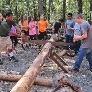 group of children learning how to use a large saw in forested area