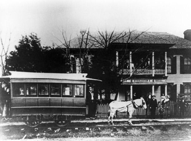 People pose outside two story building with "Huddlestons Hotel" sign on balcony and tracks with horse and streetcar