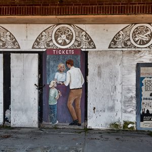 Painting on abandoned movie theater wall of white man and boy buying tickets from white woman in box office