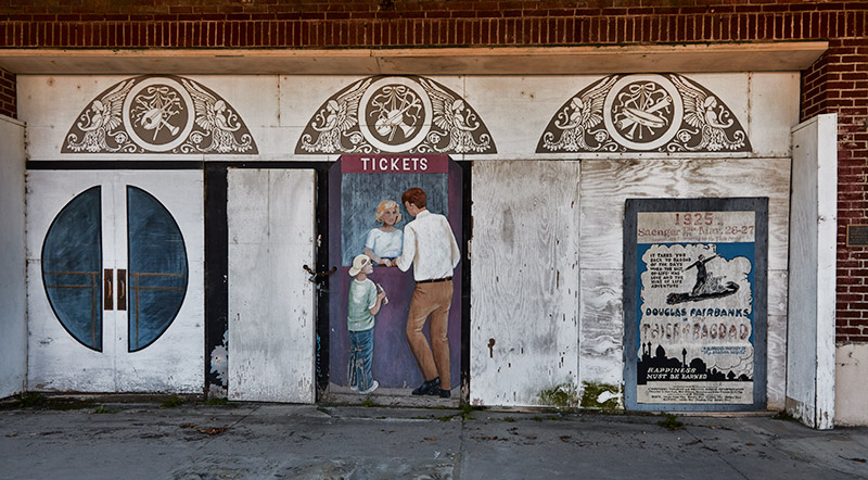 Painting on abandoned movie theater wall of white man and boy buying tickets from white woman in box office