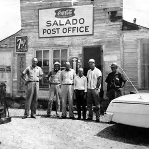 Group of white men standing outside single-story building with a car and truck