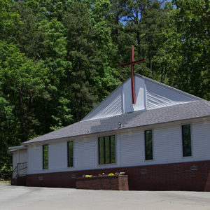 Church building with white siding and brick foundation on parking lot with sign near the road