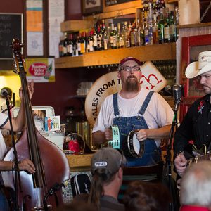 Three white musicians playing upright bass guitar and tambourine in bar with audience