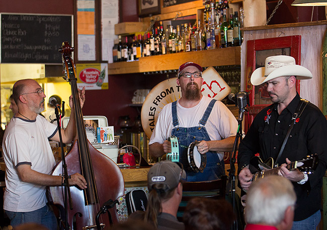 Three white musicians playing upright bass guitar and tambourine in bar with audience