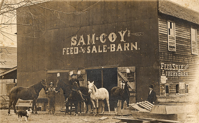White men with horses and dog standing outside "Sam Coy Feed and Sale Barn" building