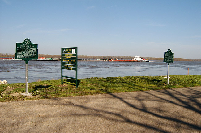 Barge on river with three signs on shore and walkway