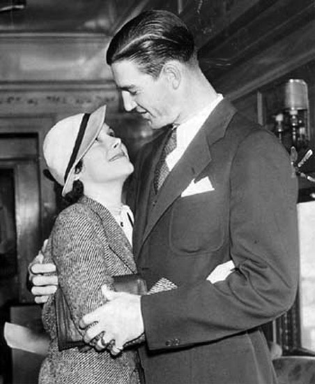 White man in suit and slicked-back hair and tie looking down at and hugging a white woman in hat who is looking up at him