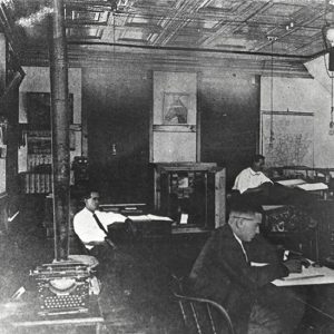 White men working at their desks in office with stove and typewriter in the foreground
