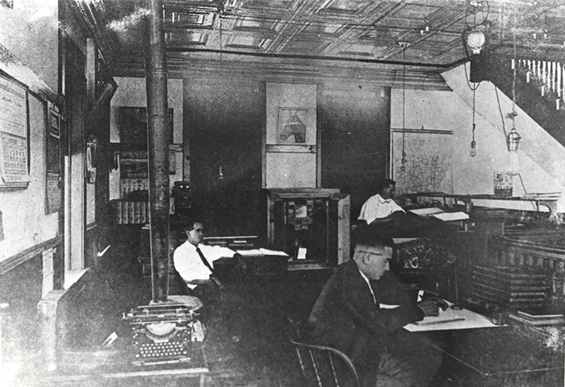 White men working at their desks in office with stove and typewriter in the foreground
