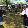 Circle of white bagpipers wearing kilts at festival with people and tents in the background
